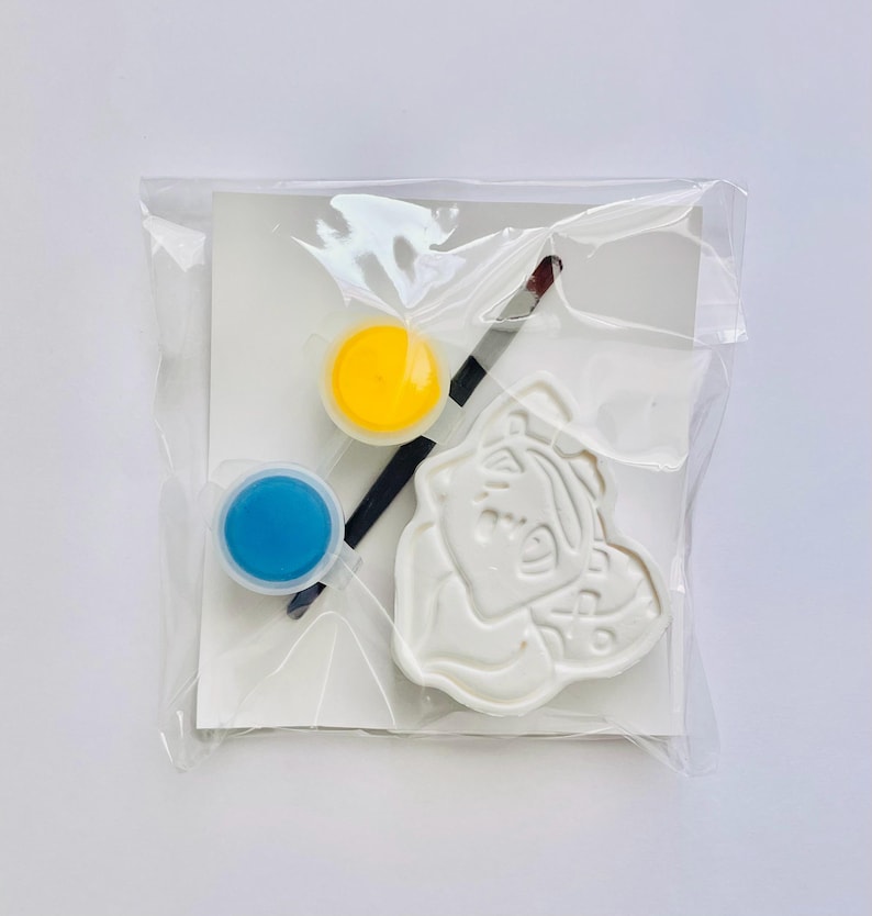 May include: A craft kit sealed in a clear plastic bag containing a white molded figure with a cartoon character design, a small paintbrush with a black handle, and two circular paint pots filled with yellow and blue paint on a white card.