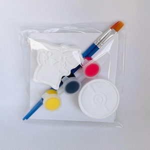May include: A craft kit in a clear plastic bag containing a white plaster figure of a character with pointed ears, two blue-handled paintbrushes, three small circular paint pots in yellow, black, and pink, and a white circular container.