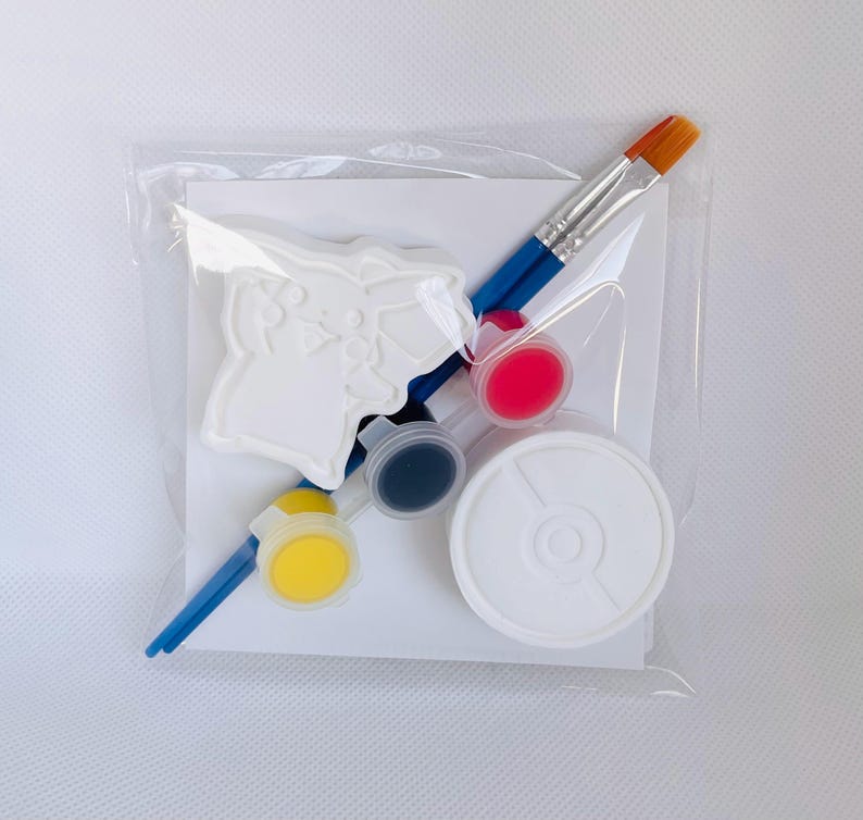 May include: A craft kit in a clear plastic bag containing a white plaster figure of a character with pointed ears, two blue-handled paintbrushes, three small circular paint pots in yellow, black, and pink, and a white circular container.