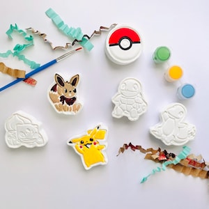 May include: A DIY craft kit featuring five white plaster figures of Pokemon characters, including Pikachu and Eevee painted in color. The set includes a blue paintbrush, three small paint pots in green, yellow, and blue, and a round red and white Pokeball.