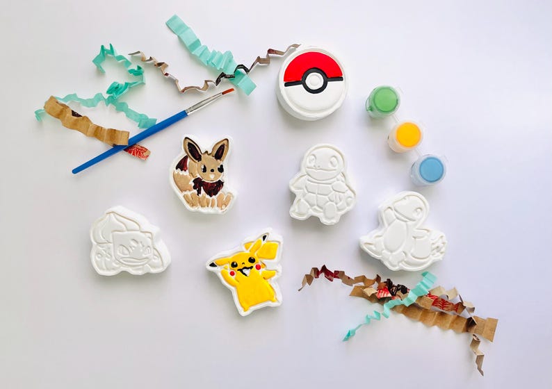 May include: A DIY craft kit featuring five white plaster figures of Pokemon characters, including Pikachu and Eevee painted in color. The set includes a blue paintbrush, three small paint pots in green, yellow, and blue, and a round red and white Pokeball.