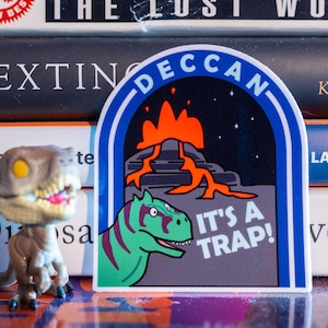 May include: A sticker with a blue arch design and the word "DECCAN" in white. The sticker features a volcano with orange lava and a green dinosaur with purple stripes. The text "IT'S A TRAP!" is also included.