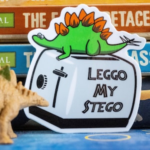 May include: A sticker featuring a green stegosaurus with orange plates atop a white toaster. The text "LEGGOMY STEGO" is printed in black. A small toy dinosaur is in the foreground.