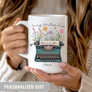 May include: White ceramic mug featuring a teal typewriter illustration with floral accents and the text "In my Writer Era." The mug also includes the name "Jane" and the phrase "writing the next chapter..."