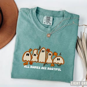 Comfort Colors® shirt with five different-shaped ghosts surrounded by pumpkins and flowers, featuring the phrase “All Bodies Are Bootiful” in bold white text