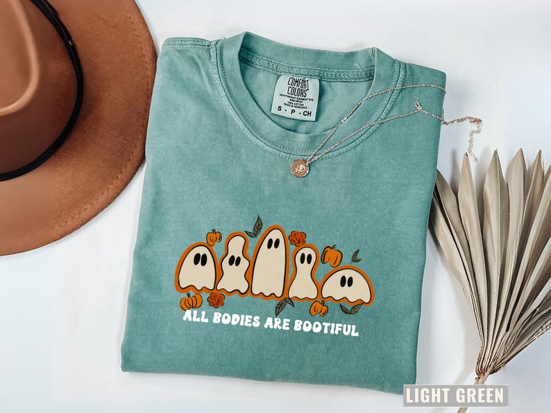 Comfort Colors® shirt with five different-shaped ghosts surrounded by pumpkins and flowers, featuring the phrase “All Bodies Are Bootiful” in bold white text