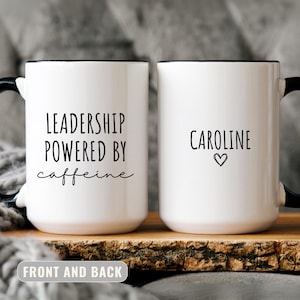 May include: Two white ceramic mugs with black handles and rims. One mug reads "LEADERSHIP POWERED BY caffeine" and the other says "CAROLINE" with a heart symbol. The mugs are on a wooden surface.