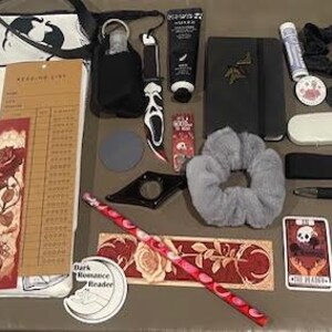 May include: A white tote bag with the words "Death By" and a skeleton holding a book. The bag is filled with various items, including a bookmark, a notebook, a pen, a hair tie, socks, and a small bag.