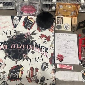 May include: A white tote bag with a black and pink design featuring the text "In My Dark Romance Era". The bag is surrounded by various items, including a book review form, a black pom-pom, a lip balm, a red and black bottle opener, and stickers.