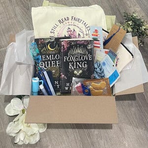May include: A gift box filled with books, a tote bag, and various accessories. The books are titled "The Emlo Queef" and "The Foxglove King." The tote bag has the text "I Still Read Fairytales" and a castle graphic. Other items include pens, eye masks, and skincare.