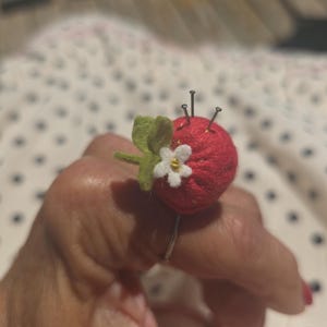 May include: A red felted strawberry pincushion ring featuring a small white flower with a yellow center and green felt leaves. The strawberry is topped with three silver sewing pins inserted into the top surface, attached to a simple metal ring band.