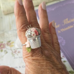May include: A silver thimble ring with a floral pin cushion top. The ring features a small pink flower and green leaf accent. Three pins are inserted into the cushion. The ring is worn on a finger.
