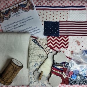 May include: A craft kit featuring a collection of fabric scraps with red, white, and blue patterns, a wooden spool, spools of thread, a small bag of assorted buttons and sequins, and an informational card titled Patriotic Snippet Spool Kit.