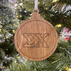 Può includere: Ornamento natalizio in legno di forma rotonda e finitura marrone chiaro. L'ornamento presenta le lettere greche Σ e X in un design ritagliato. Un nastro bianco è attaccato per appenderlo. L'ornamento è esposto su un albero di Natale.