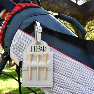 Könnte beinhalten: Eine Golftasche mit einem weißen Teehalter aus Leder. Der Halter hat sechs Golf-Tees aus Holz und die Buchstaben "ПВФ" aufgedruckt. Die Golftasche ist blau, rot und weiß.