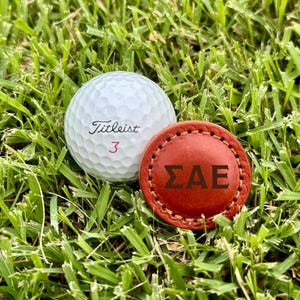 May include: A white Titleist golf ball with the number 3 next to a round, red leather golf ball marker with the Greek letters ΣAE. The items are resting on a bed of green grass.