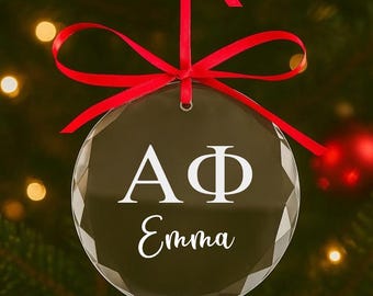 Puede incluir: Un adorno navideño redondo y transparente con un diseño facetado, colgado de una cinta roja. El adorno presenta las letras griegas ΑΦ y el nombre "Emma" en escritura blanca, sobre un fondo oscuro.
