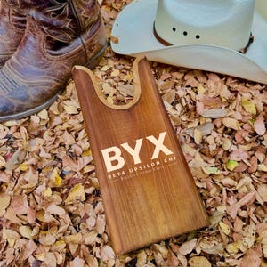 以下が含まれることがあります： 「BYX BETA UPSILON CHI Brothers Under Christ」という文字が刻まれた木製ブーツジャック。ブーツジャックは落ち葉の上に置かれています。
