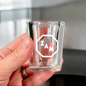 May include: A clear glass shot glass with a white octagon design that says "ΣΑΜ" on it.