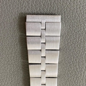 Peut inclure: Gros plan sur un bracelet de montre argenté. Le bracelet est composé de maillons métalliques rectangulaires avec une finition brossée. Les maillons sont reliés selon un motif répétitif. Le bracelet est sur une surface grise texturée.