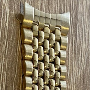 May include: Close-up of a gold-colored watch band. The band is made of small, rectangular links that are connected to each other. The band has a curved end piece and a clasp.