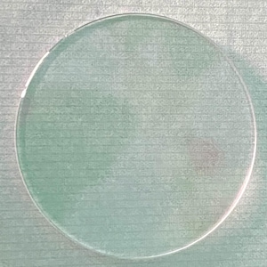 May include: A circular, transparent glass disc with a smooth, polished surface and clear edges. The object is flat and uniform in shape, featuring a neutral, colorless appearance that allows light to pass through it completely without any distortion.