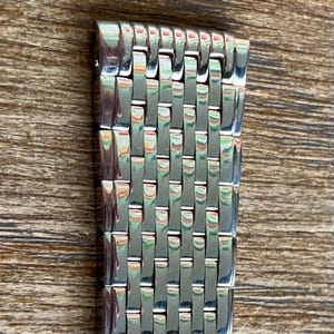 May include: Close-up of a silver-toned watch band with a woven design. The band is made of small, rectangular links that are interlocked to create a flexible strap. The band is shown against a wooden background.