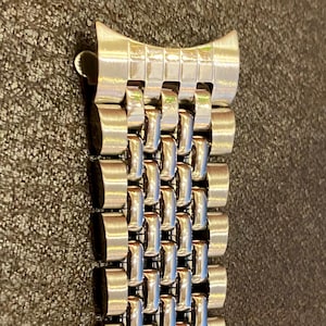 May include: A close-up of a silver-colored watch band. The band is made of small, rectangular metal links that are connected together. The band has a curved end piece with a clasp.