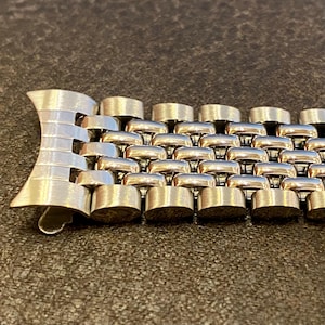 May include: Close-up of a silver-toned metal bracelet. The bracelet features a unique design with interconnected, cylindrical links. The clasp is a stylized, curved shape. The bracelet is displayed on a dark, textured surface.