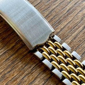 May include: Close-up of a two-tone watch band. The band features a brushed silver clasp and a woven pattern of silver and gold-colored links. The band is set against a wooden surface.