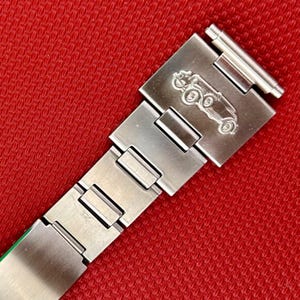 May include: A silver-toned metal watch bracelet featuring a brushed finish and a rectangular clasp. The clasp is engraved with a detailed, raised relief design of a vintage automobile, showing wheels and a chassis, positioned above the link structure.