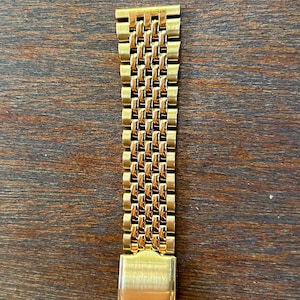 May include: A gold-toned metal watch band featuring a multi-link design with a polished finish. The strap consists of several rows of small, rectangular interlocking links that create a textured appearance, ending in a smooth, solid metal clasp.