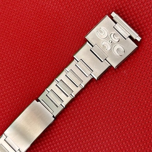 May include: A silver-toned metal watch bracelet featuring a brushed finish and a series of rectangular links. The end link displays a distinct embossed logo consisting of three crescent moon shapes arranged around a central circular dot.