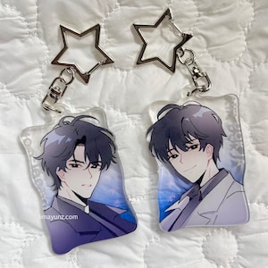 May include: Two clear acrylic keychains with anime-style illustrations of a person with dark hair and a white suit. Each keychain has a silver star-shaped top and a clasp. The website limayunz.com is visible on one keychain.