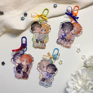 May include: Four clear acrylic keychains with colourful ribbons and metal clasps. Each keychain features a pair of cartoon characters, one with brown hair and the other with blonde hair. The characters are holding each other and smiling.