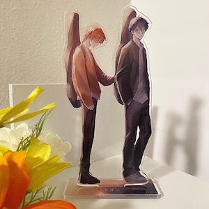 Puede incluir: Dos personajes de anime se encuentran uno al lado del otro. El personaje de la izquierda tiene el pelo naranja y lleva una chaqueta marrón. El personaje de la derecha tiene el pelo negro y lleva una chaqueta negra. Ambos personajes sostienen guitarras. La base del soporte tiene la palabra "サワリ" escrita en ella.