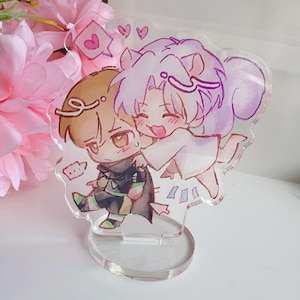 May include: A clear acrylic stand with a colourful illustration of two anime-style characters embracing. One character has brown hair and the other has purple hair. The stand has a circular base and a decorative shape.