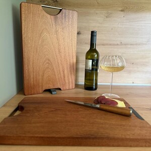 Op de afbeelding: Twee houten snijplanken, één rechtop met een metalen handvat en één plat met een mes en kaas. Een wijnfles en een wijnglas zijn ook aanwezig. De planken zijn gemaakt van roodbruin hout.
