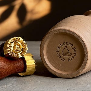 May include: A terracotta-colored pottery vase with the text "matcha house pottery" stamped on the bottom. A wooden handled stamp with a gold-colored design is next to the vase, along with two pottery tools.