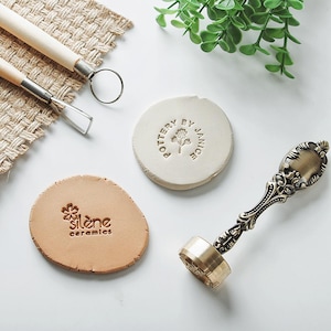 Puede incluir: Una colección de herramientas y materiales de cerámica, que incluye un sello de metal con un mango adornado, dos herramientas de arcilla y dos piezas de arcilla con diseños estampados. Una pieza de arcilla está estampada con "Pottery by Janice", y la otra con "silène ceramics".