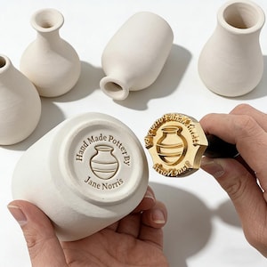 Puede incluir: Una colección de pequeños jarrones de cerámica sin esmaltar de varias formas. Se sostiene un jarrón, mostrando el texto estampado "Hand Made Pottery By Jane Norris". También es visible un sello de madera con el mismo logotipo.