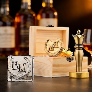 Puede incluir: Un conjunto de accesorios de bar, que incluye un cubito de hielo plateado con las iniciales "BM", un tapón de botella dorado y una caja de madera con un emblema dorado. Un tapón de botella dorado con forma de pieza de ajedrez está de pie. El fondo presenta botellas de licor.