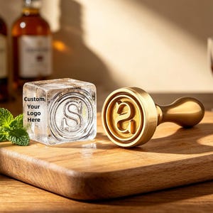 May include: A custom ice cube stamp and an ice cube with the letter "S" inside. The stamp is made of gold-colored metal and has a handle. The ice cube has the text "Custom Your Logo Here" on it. The items are on a wooden cutting board.