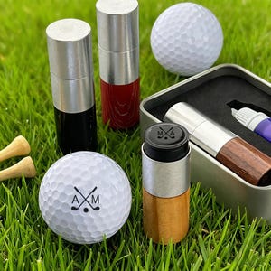 May include: Golf-themed accessories on green grass. Includes golf balls with initials and a logo, cylindrical containers with silver tops, and wooden tees. Some containers are black, red, and wood-toned. A metal box holds a pen and a container.