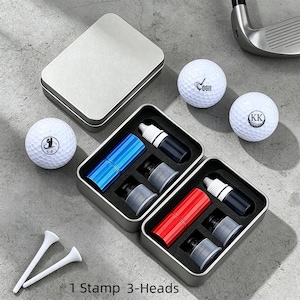 May include: A golf accessory set featuring two metal tins with blue and red stamp pens, ink bottles, and golf ball stamps. Includes two white golf tees and three golf balls. The text "1 Stamp 3-Heads" is visible.