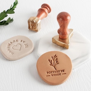 May include: A collection of handmade pottery stamps with wooden handles and brass stamp heads. One stamp reads "HANDMADE BY JIL" with a heart, and another says "POTTERY BY WENDY" with a botanical design. The stamps are in various shapes and sizes.