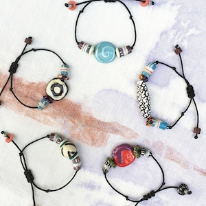 May include: Five handmade bracelets with black cords and colorful ceramic beads. The beads are various shapes and sizes, including round, oval, and cylindrical, with patterns in blue, white, red, and black. The bracelets are displayed on a white and blue patterned surface.