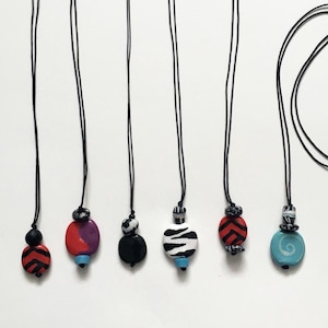 May include: Six handmade pendant necklaces with black cords. Each necklace features a unique, colorful bead design. The beads include patterns like red and black chevrons, zebra stripes, and swirls. The background is a plain white surface.
