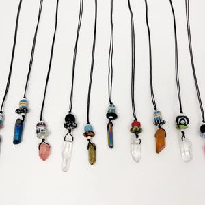 May include: A collection of handmade necklaces with black cords and various crystal pendants. Each necklace features a unique combination of beads in different colors and shapes, with crystal pendants in shades of gray, pink, clear, and orange.