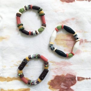 May include: Three colorful beaded bracelets with different patterns and colors. The bracelets are made with various shapes and sizes of beads, including round, oval, and rectangular beads. The beads are in shades of brown, green, red, white, and black.